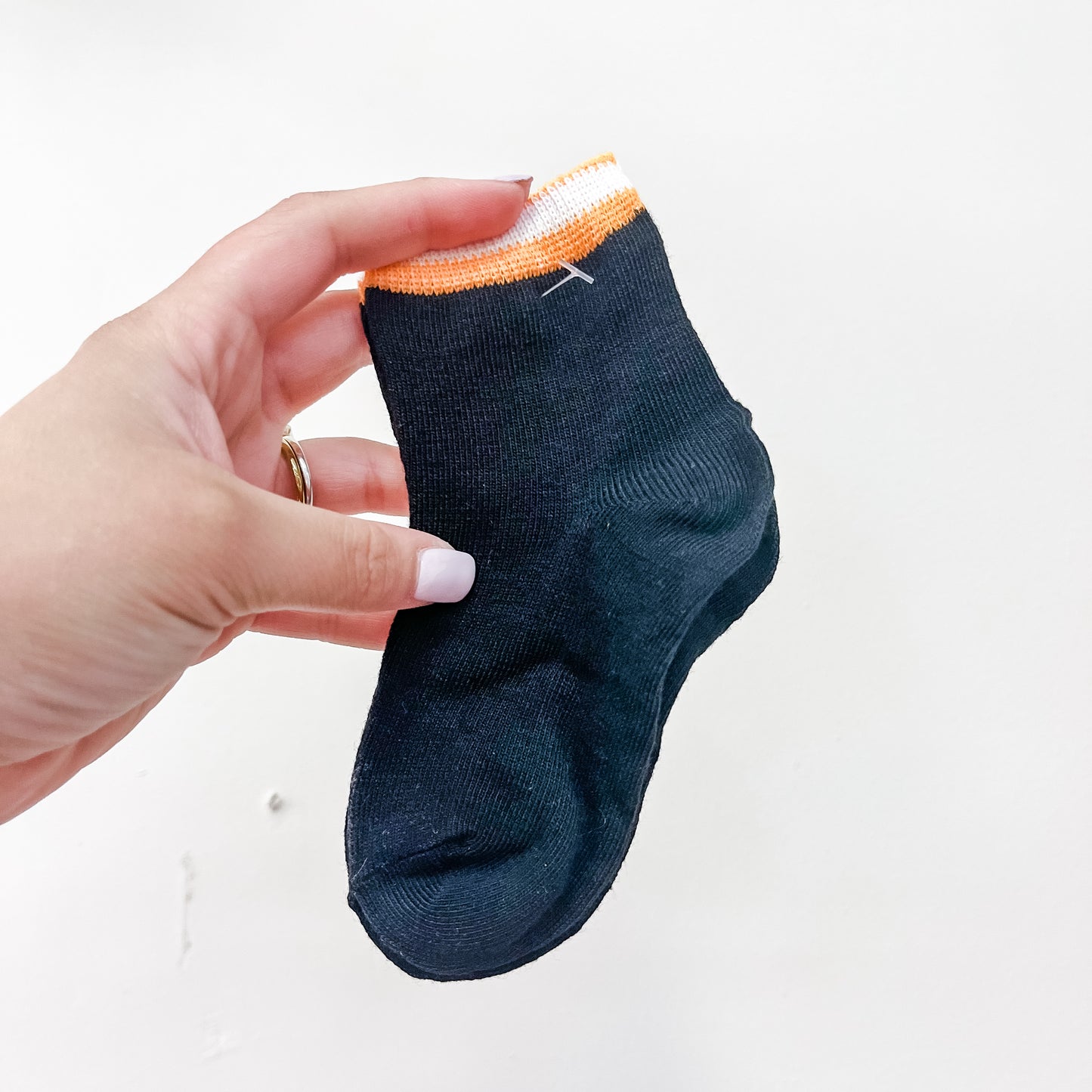 A hand with light pink nail polish holds a pair of Boo Crew Socks by Tiny Tot Bundles—small black socks featuring an orange and white stripe at the top—against a plain white background.