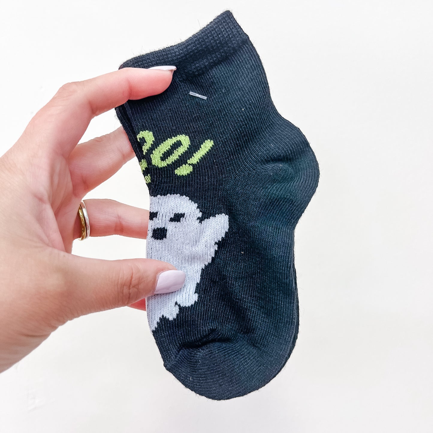 A hand with light skin, neatly manicured nails, and a gold ring on the ring finger holds a pair of Tiny Tot Bundles Boo Crew Socks—a small black sock featuring a white ghost and green text—against a plain white background.