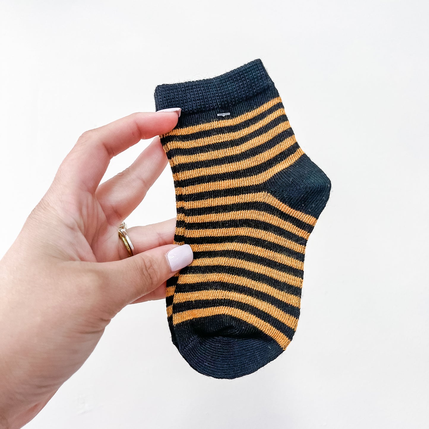 A hand with light pink nail polish holds a Tiny Tot Bundles Boo Crew Sock, featuring black and orange stripes, against a plain white background.