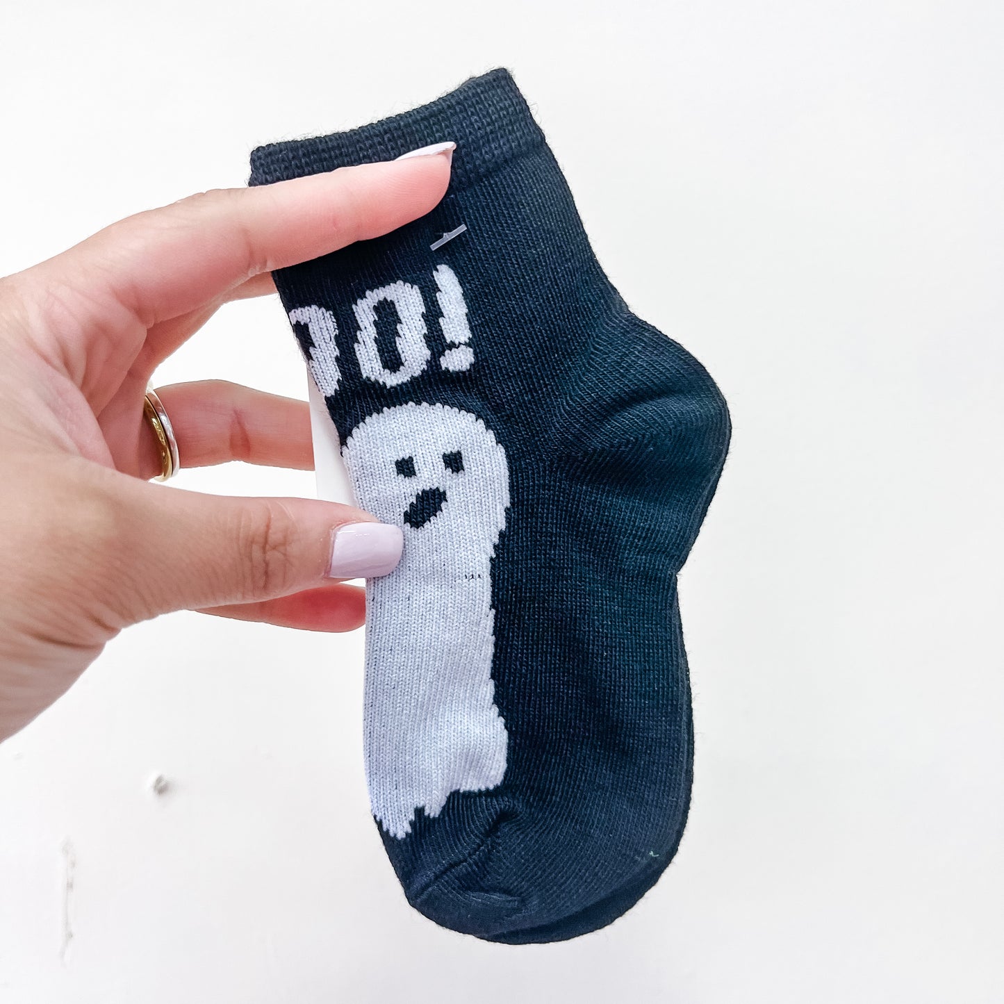 A hand with light purple nail polish holds Tiny Tot Bundles’ Boo Crew Socks—a small black sock featuring a white ghost and the word “BOO!”—against a white background.