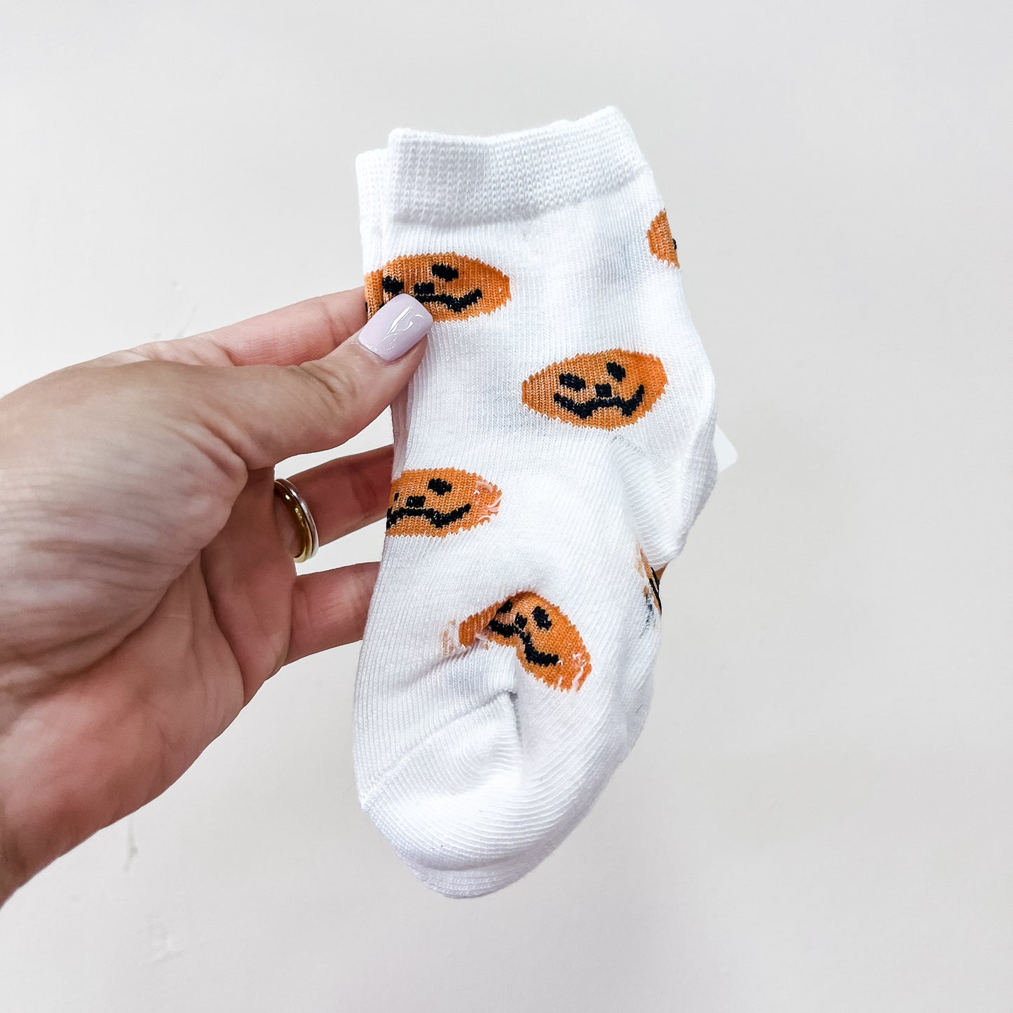 A hand holds a Tiny Tot Bundles Boo Crew Sock—a small white sock adorned with orange jack-o-lantern faces—against a plain white background.