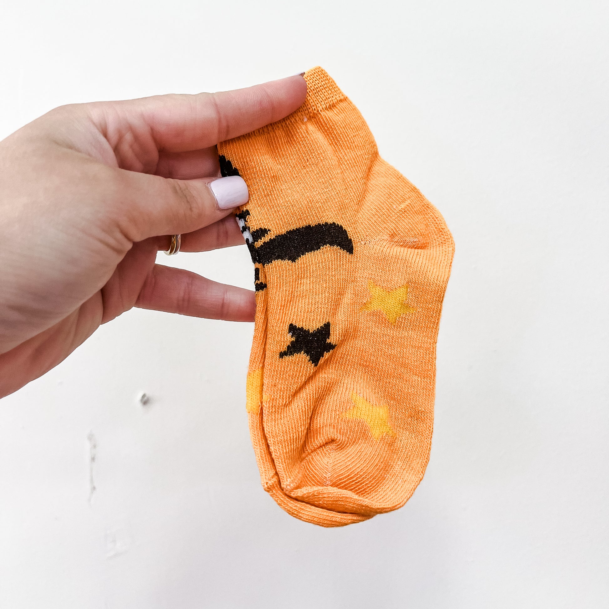 A hand with light pink nail polish holds a pair of Tiny Tot Bundles Boo Crew Socks—small orange socks featuring black and yellow designs—against a plain white background.