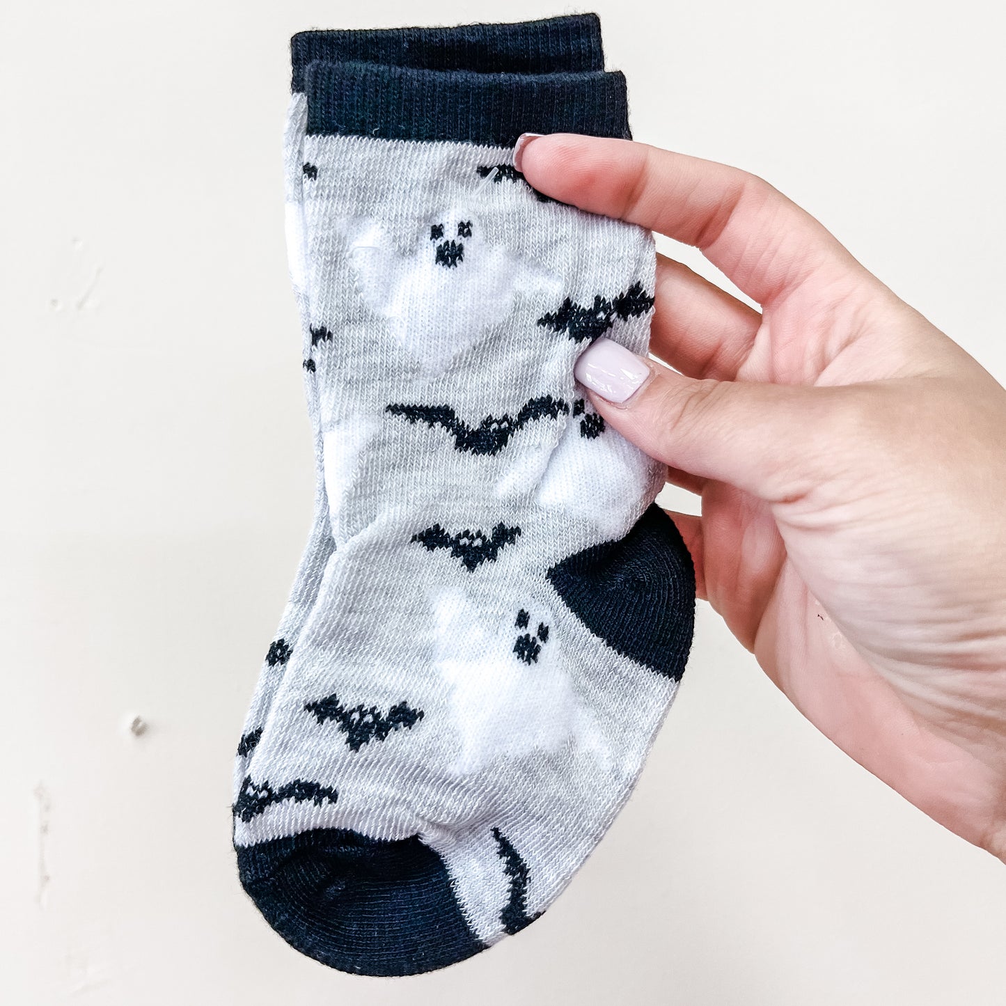 A hand holds a pair of Tiny Tot Bundles Spooky Steps Socks, featuring small gray fabric adorned with black bats and ghost patterns. The socks have dark cuffs, heels, and toes, set against a plain light background.