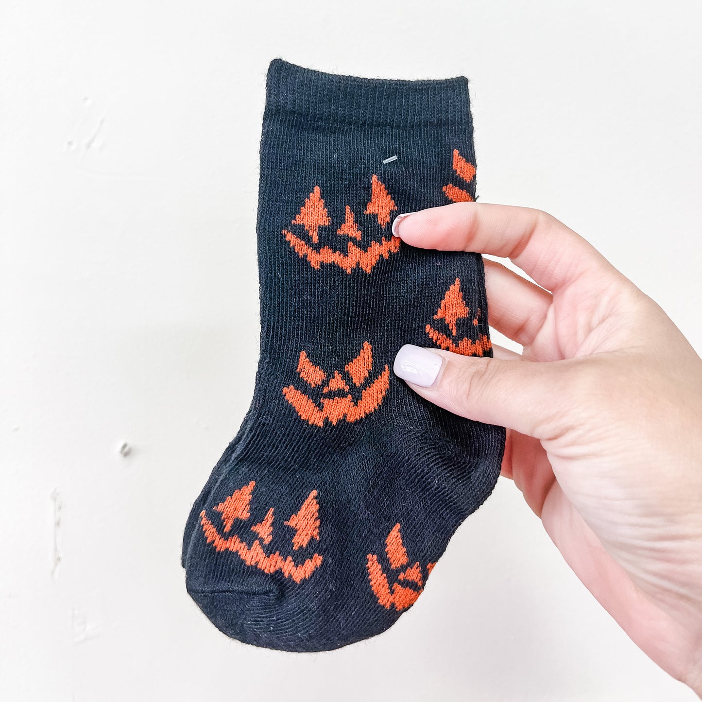 A hand holds the Spooky Steps Socks by Tiny Tot Bundles—small black socks adorned with orange jack-o-lantern faces—against a plain white background.