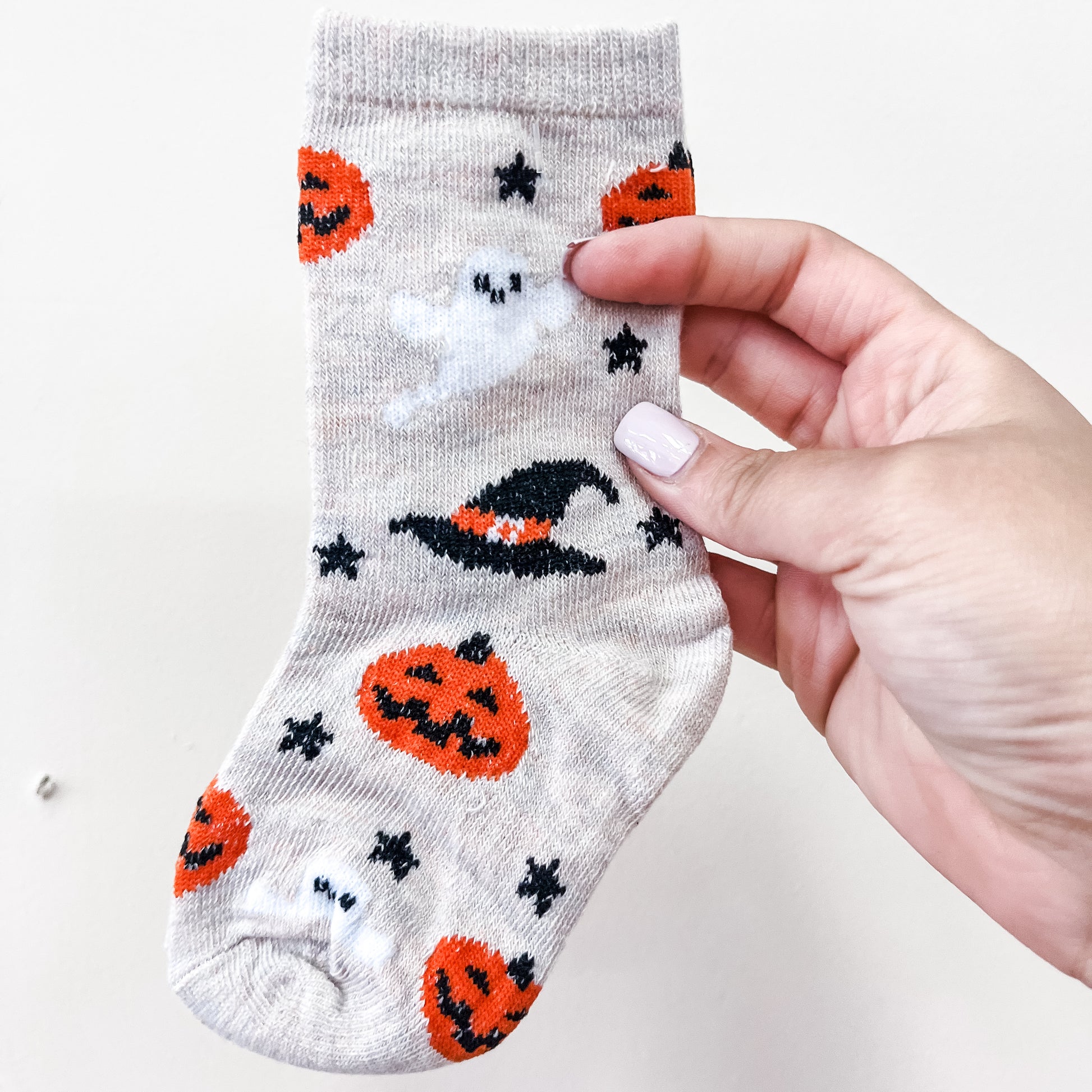 A hand holds a pair of Spooky Steps Socks by Tiny Tot Bundles, beige in color and decorated with Halloween-themed designs such as jack-o-lanterns, a witch hat, ghosts, and black stars.