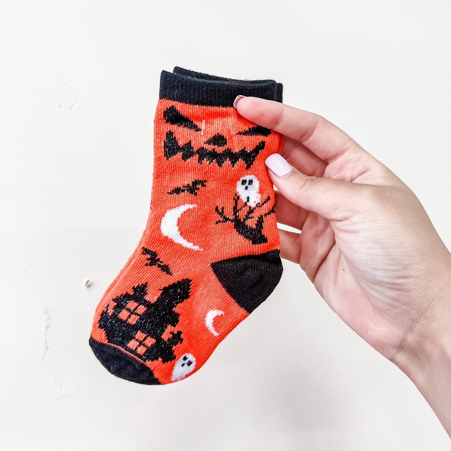 A hand holds a Spooky Steps Sock by Tiny Tot Bundles, a small orange sock with black trim featuring Halloween designs such as a haunted house, ghosts, bats, a crescent moon, and the word “Mew” at the top.