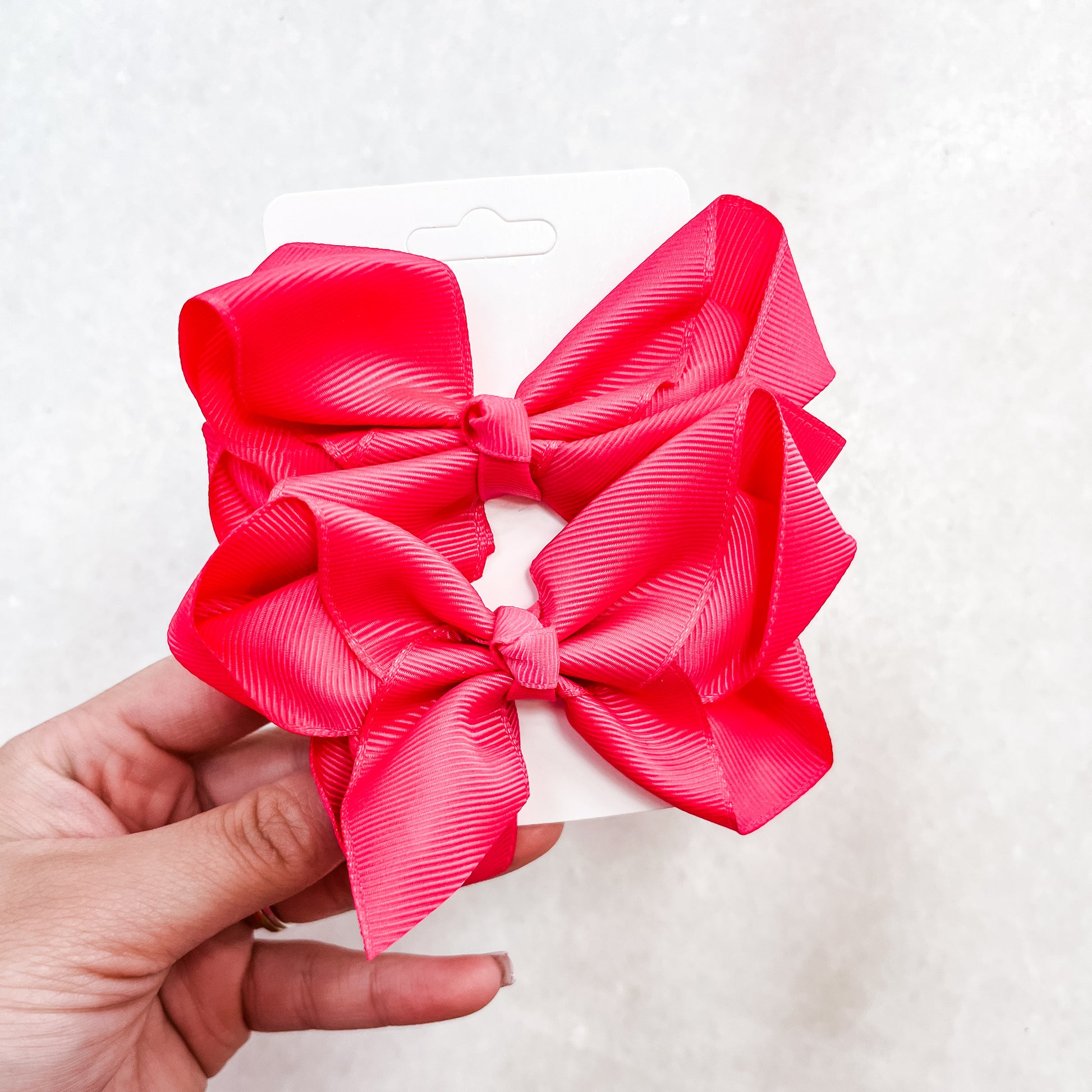 A hand holds a white card displaying the Pretty In Pink Bow Set by Tiny Tot Bundles, featuring two large bright pink hair bows against a light textured background.