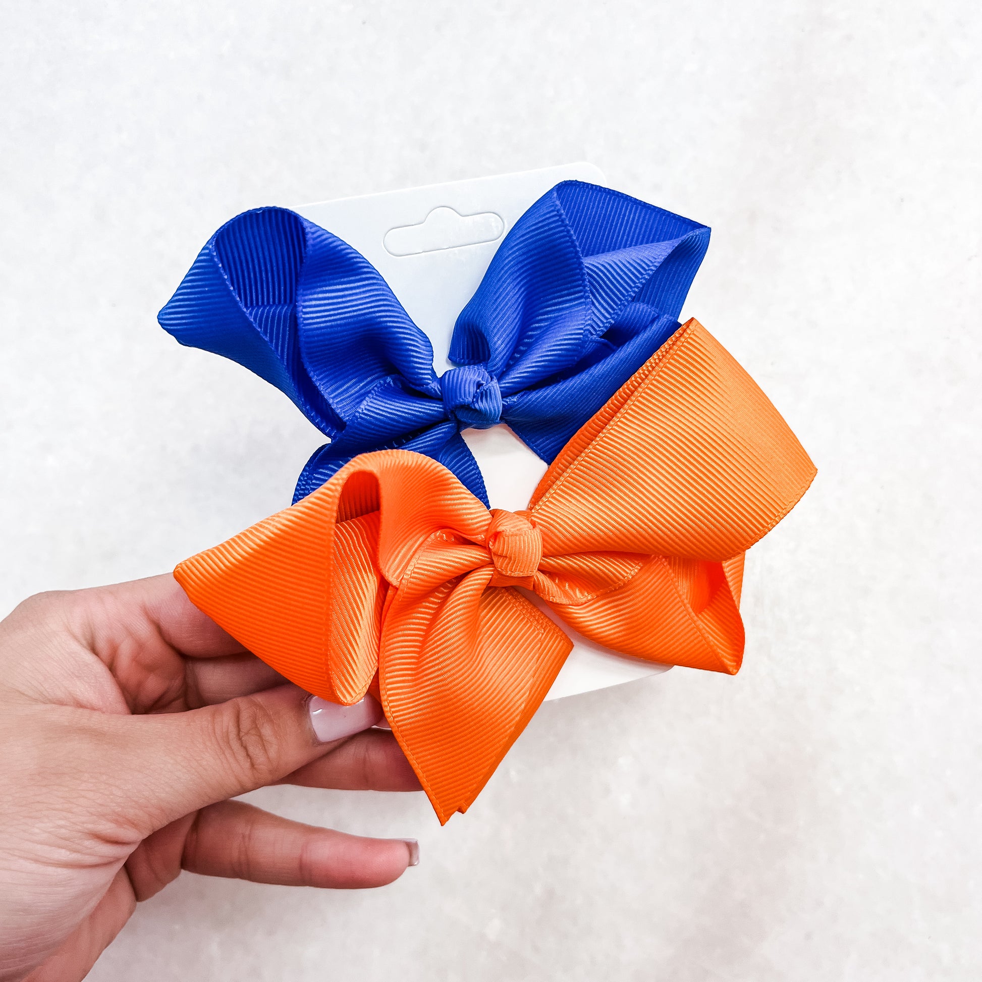 A hand holds a white card featuring the Tiny Tot Bundles Gator Spirit Bow Set: two large textured hair bows, one royal blue and one bright orange, against a light neutral background.
