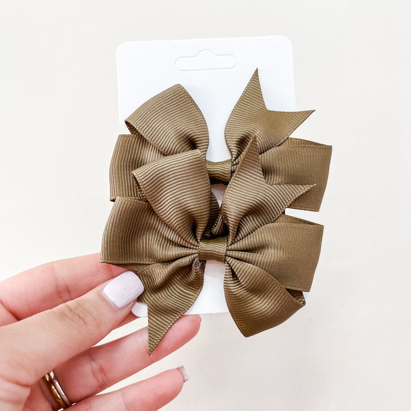 A hand with pale nail polish holds a white card displaying two large olive-green ribbed fabric bows from the Everyday Essentials Bow Set by Tiny Tot Bundles against a light background.