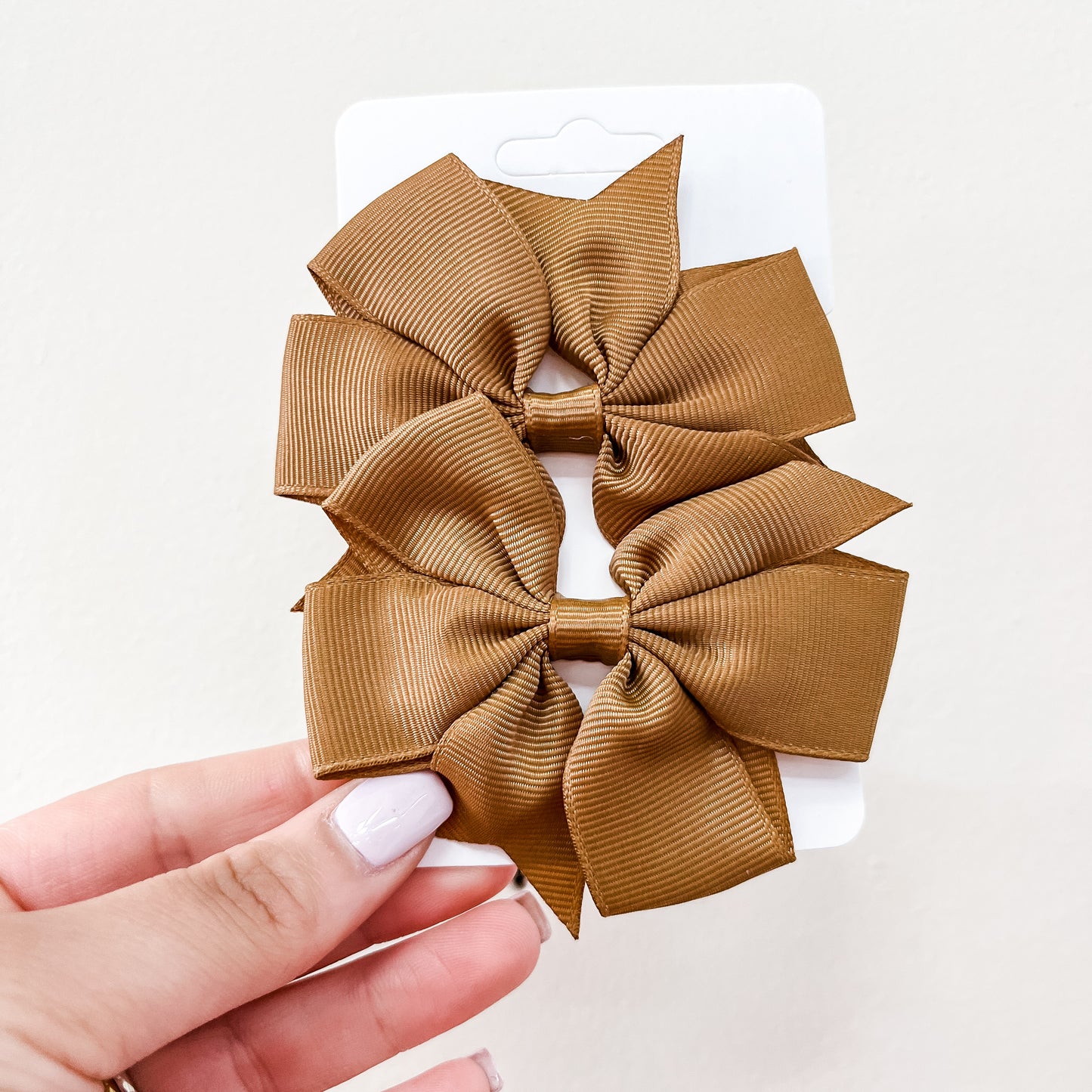 A hand with light pink nail polish holds a white card displaying two brown, ribbed fabric bows from the Everyday Essentials Bow Set by Tiny Tot Bundles against a plain light background.