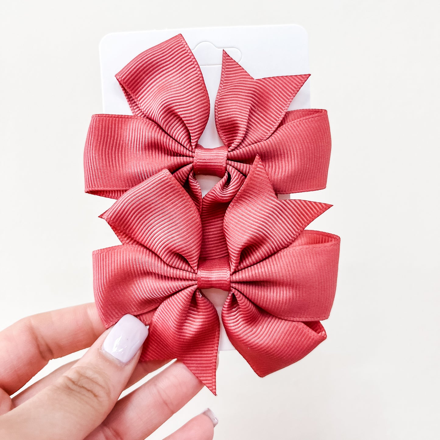 A hand with light purple nail polish holds a card featuring two large, textured pink bows from the Everyday Essentials Bow Set by Tiny Tot Bundles against a plain white background.
