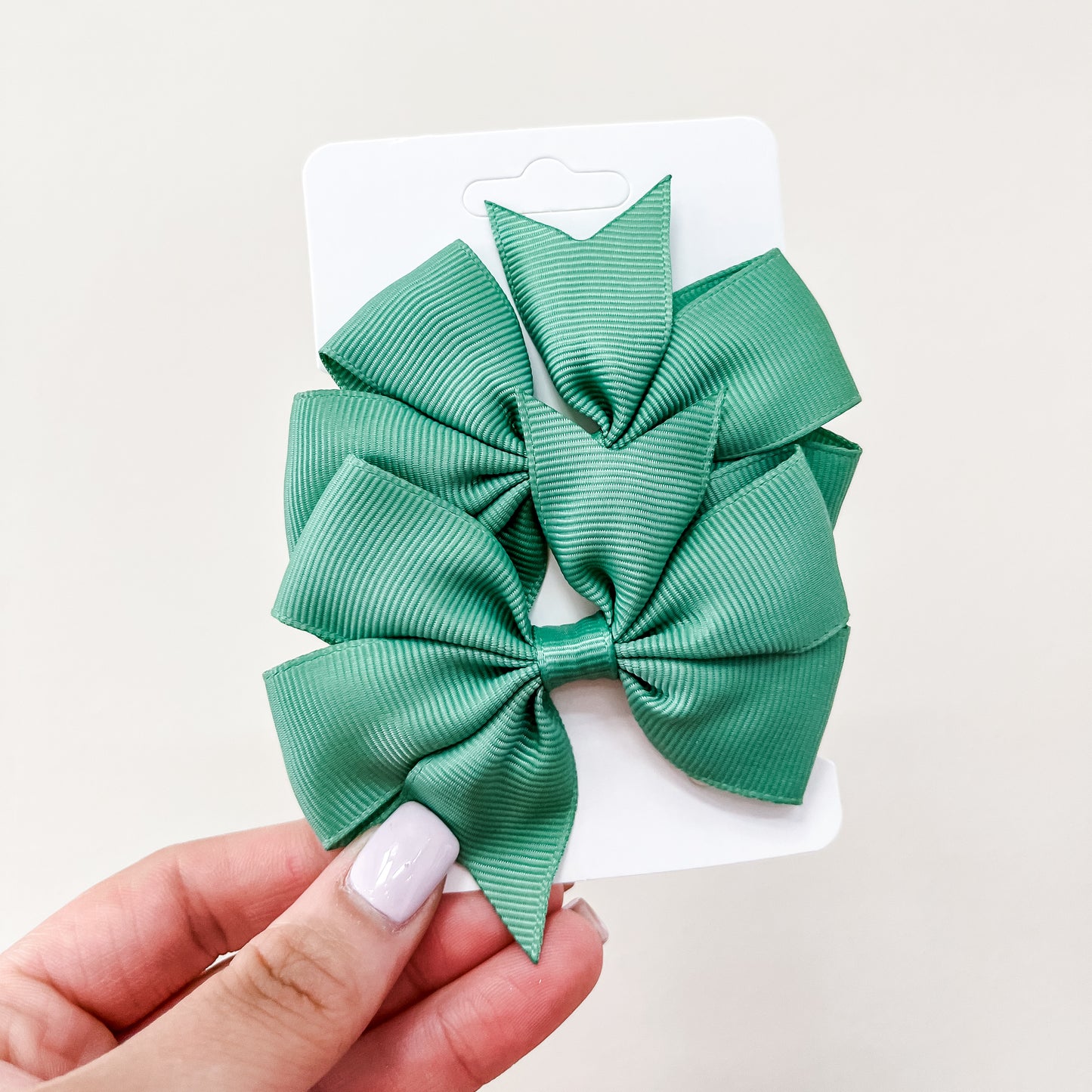 A hand with light nail polish holds a white card featuring two large, green pointed ribbon bows from the Everyday Essentials Bow Set by Tiny Tot Bundles against a plain, light background.