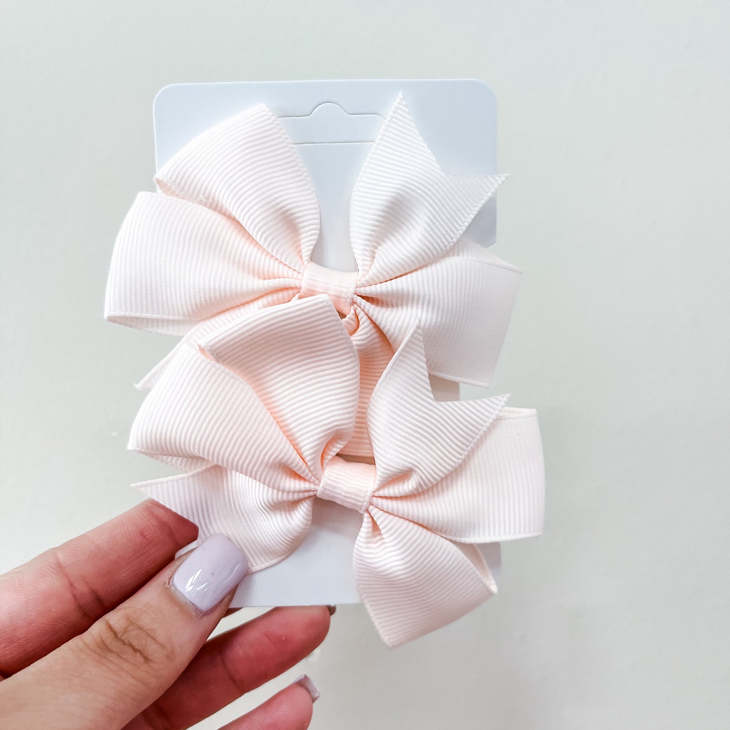 A hand holds a white card featuring two pale pink, ribbed hair bows from the Everyday Essentials Bow Set by Tiny Tot Bundles, set against a plain light background.