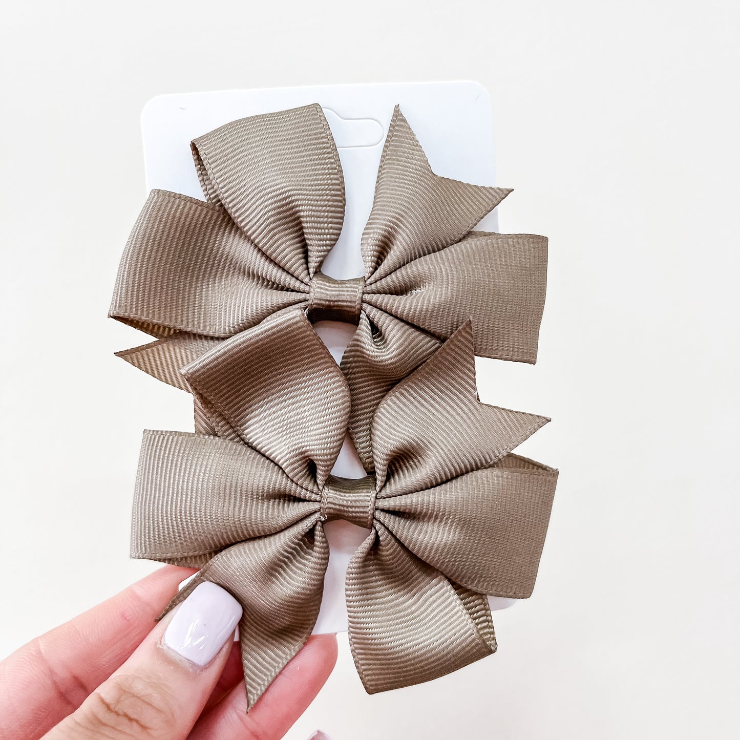 A hand holds a white card displaying two matching taupe hair bows from the Everyday Essentials Bow Set by Tiny Tot Bundles, set against a plain light-colored background.