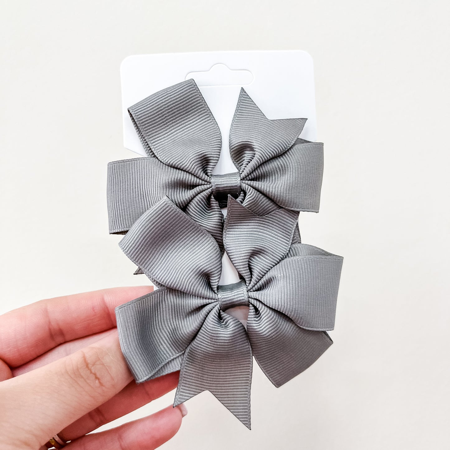 A hand holds a white card with two large gray grosgrain bows from the Tiny Tot Bundles Everyday Essentials Bow Set, shown vertically against a plain white background.