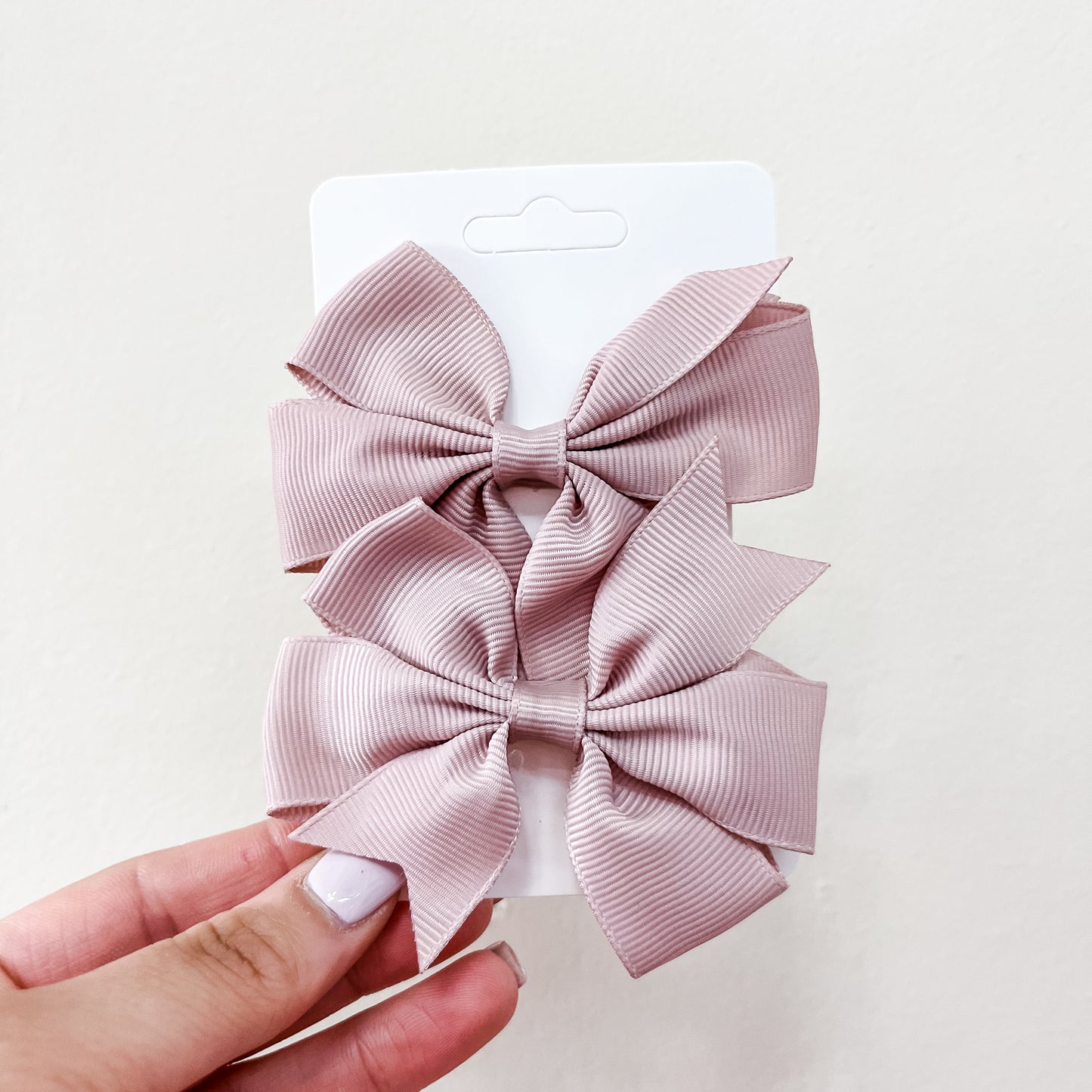 A hand holds a white card with two large, light pink, ribbed fabric bows from the Everyday Essentials Bow Set by Tiny Tot Bundles, set against a plain cream background.