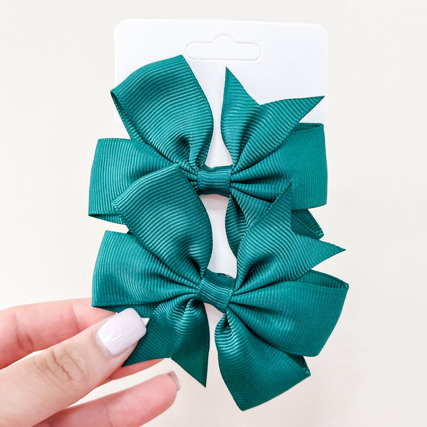 A hand with light nail polish holds a white card featuring the Everyday Essentials Bow Set by Tiny Tot Bundles—two large, ribbed teal hair bows—shown against a pale background.
