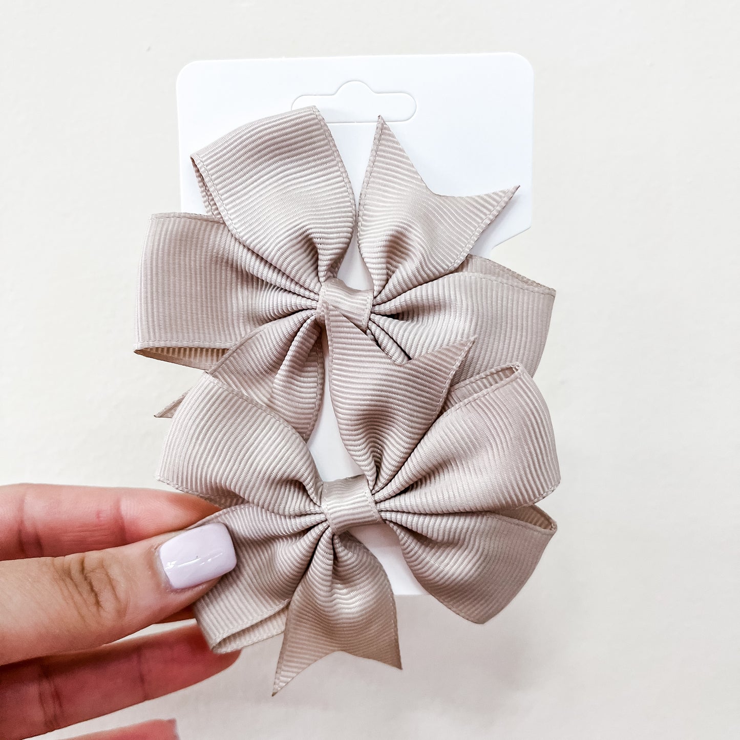 A hand with light pink nails holds a white card featuring two large, beige, ribbed hair bows from the Tiny Tot Bundles Everyday Essentials Bow Set against a pale background.