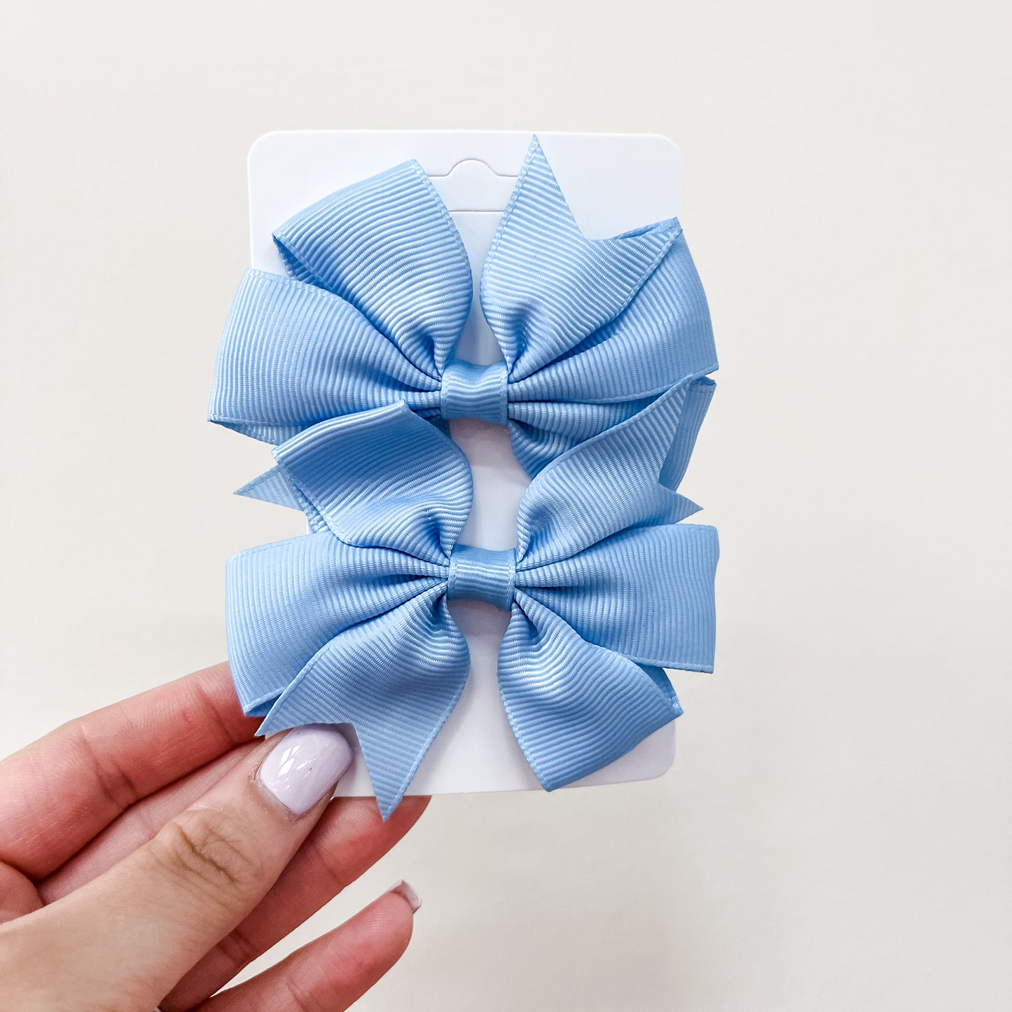A hand with light purple nail polish holds a white card featuring the Tiny Tot Bundles Everyday Essentials Bow Set, displaying two large, neatly tied light blue bows against a plain background.
