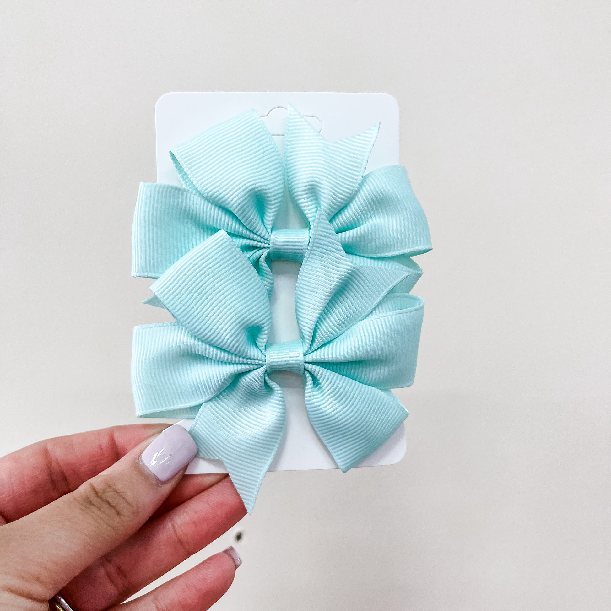 A hand with light purple nail polish holds a card featuring two large, light blue bows from the Everyday Essentials Bow Set by Tiny Tot Bundles, displayed against a plain, light background.