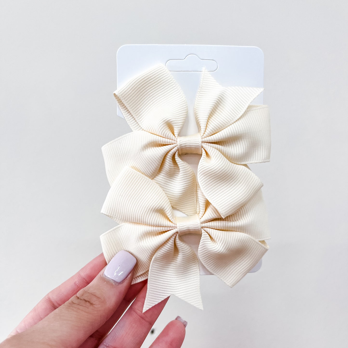 A hand with light purple nails holds a card displaying two large, cream-colored ribbed fabric bows from the Everyday Essentials Bow Set by Tiny Tot Bundles against a plain light background.
