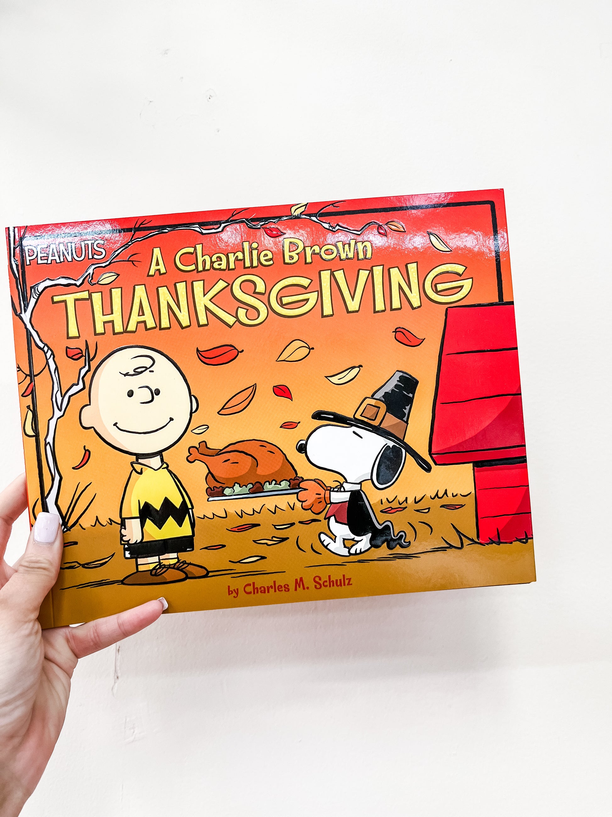 A hand holds the Simon & Schuster "A Charlie Brown Thanksgiving Book," showing Charlie Brown and Snoopy in a pilgrim hat with a turkey, autumn leaves, and the red doghouse in the background.