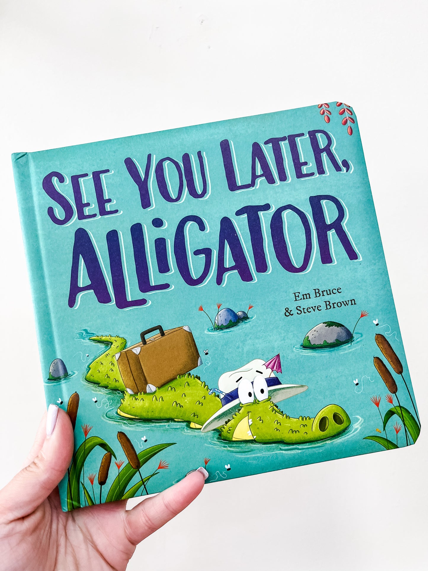 A hand holds the See You Later, Alligator Book by Simon & Schuster, featuring a cartoon alligator with a suitcase on the cover, surrounded by reeds and small rocks in water.