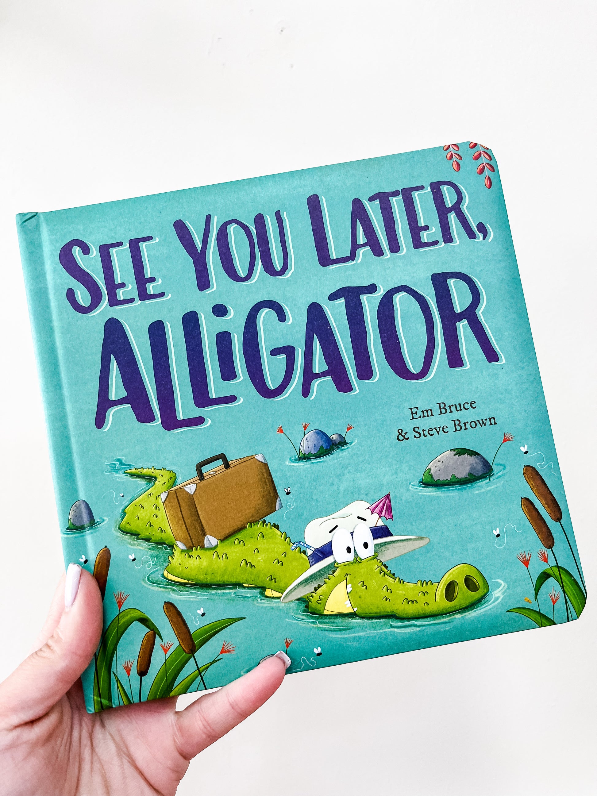 A hand holds the See You Later, Alligator Book by Simon & Schuster, featuring a cartoon alligator with a suitcase on the cover, surrounded by reeds and small rocks in water.