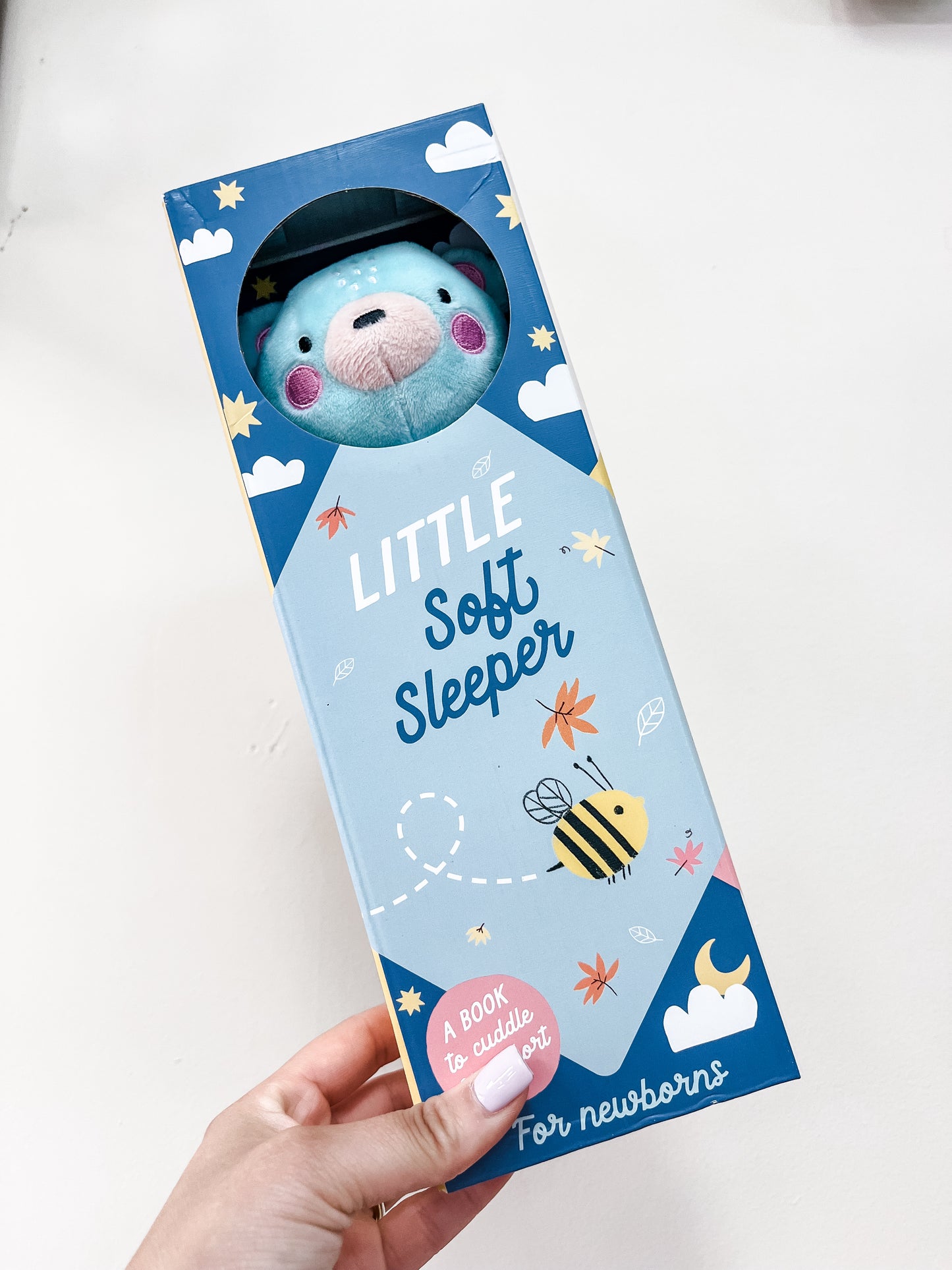 A hand holds a Simon & Schuster Little Soft Sleeper gift set for newborns, with a blue plush bear visible through a circular window and packaging decorated with leaves, hearts, and a bee.