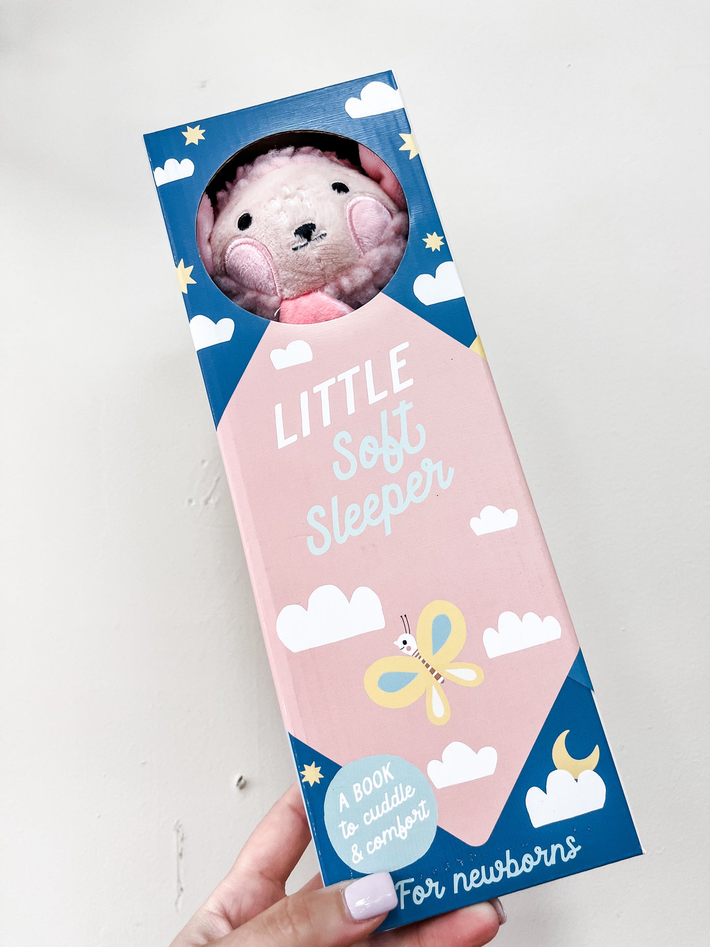 A hand holds a boxed Simon & Schuster "Little Soft Sleeper" gift set featuring a plush pink bear. The packaging displays clouds, stars, and a butterfly, and reads: "A book to cuddle & comfort for newborns.
