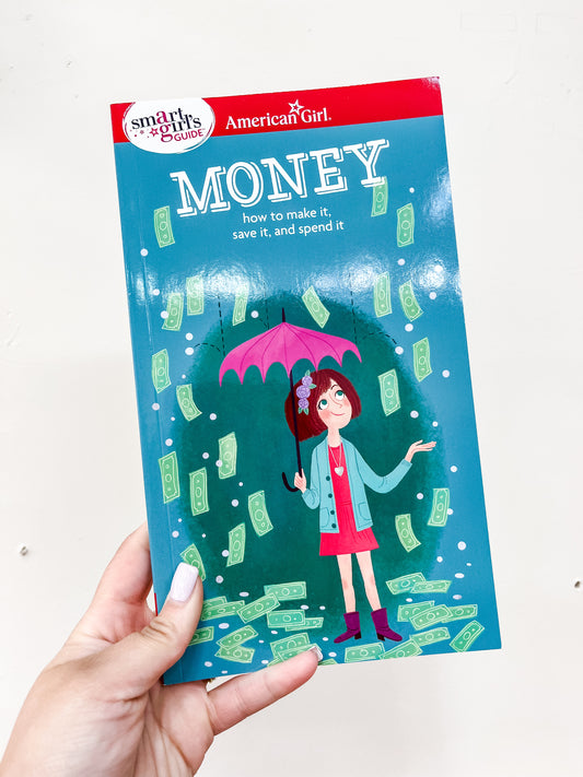 A hand holds the Smart Girl's Guide - Money Book from Simon & Schuster. The cover features a girl with an umbrella surrounded by falling dollar bills.
