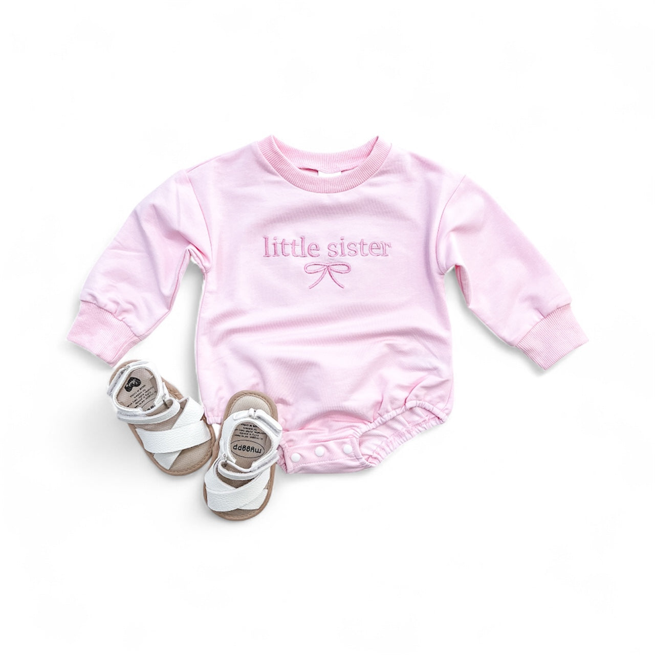 The Tiny Tot Bundles Little Sister Bow Bubble, a pink long-sleeve baby onesie with "little sister" and a bow on the front, is shown next to white baby sandals on a white background.