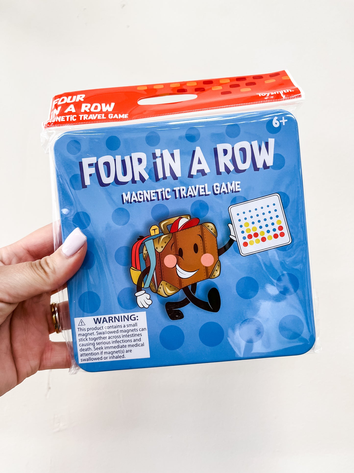 A hand holds a blue tin with a red top labeled Toy Smith Magnetic Travel Game - Four In A Row for ages 6+, featuring a smiling cartoon suitcase holding the game board and a warning label at the bottom.