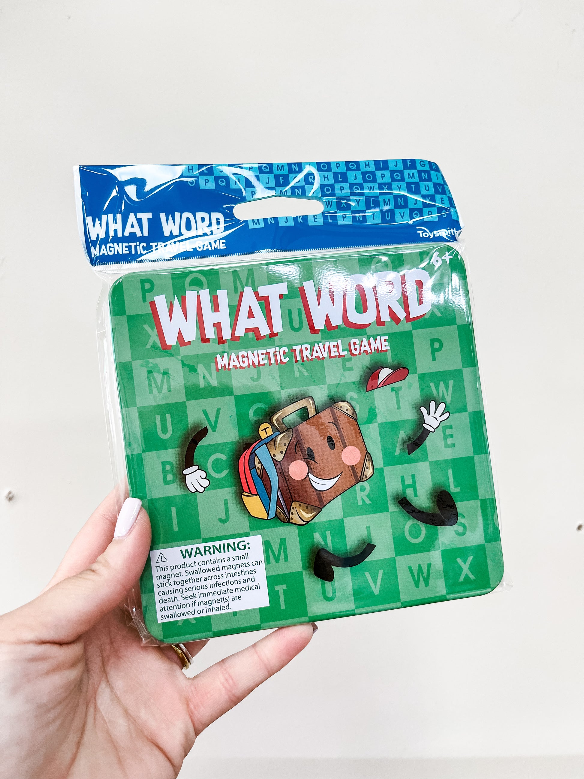 A hand holds a green Toy Smith Magnetic Travel Game – What Word tin, featuring a smiling suitcase cartoon, black letters A–Z, a blue top, and a warning label.