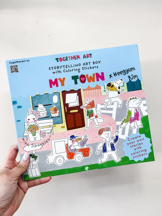 A hand holds a colorful box labeled "Storytelling Art Box - My Town" by Together Art, featuring illustrated animal characters and scenes on the cover.