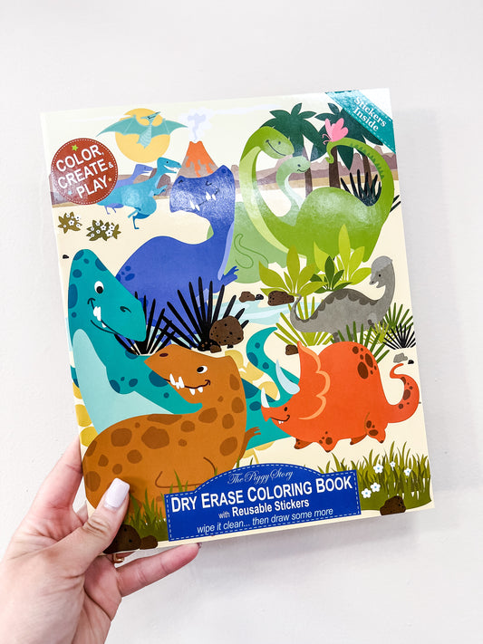 A hand holds The Piggy Store’s Dinosaur Around the World Dry Erase Coloring Book, which features colorful cartoon dinosaurs, a "Color, Create, Play" sticker, and a label for reusable stickers and wipe-clean pages.