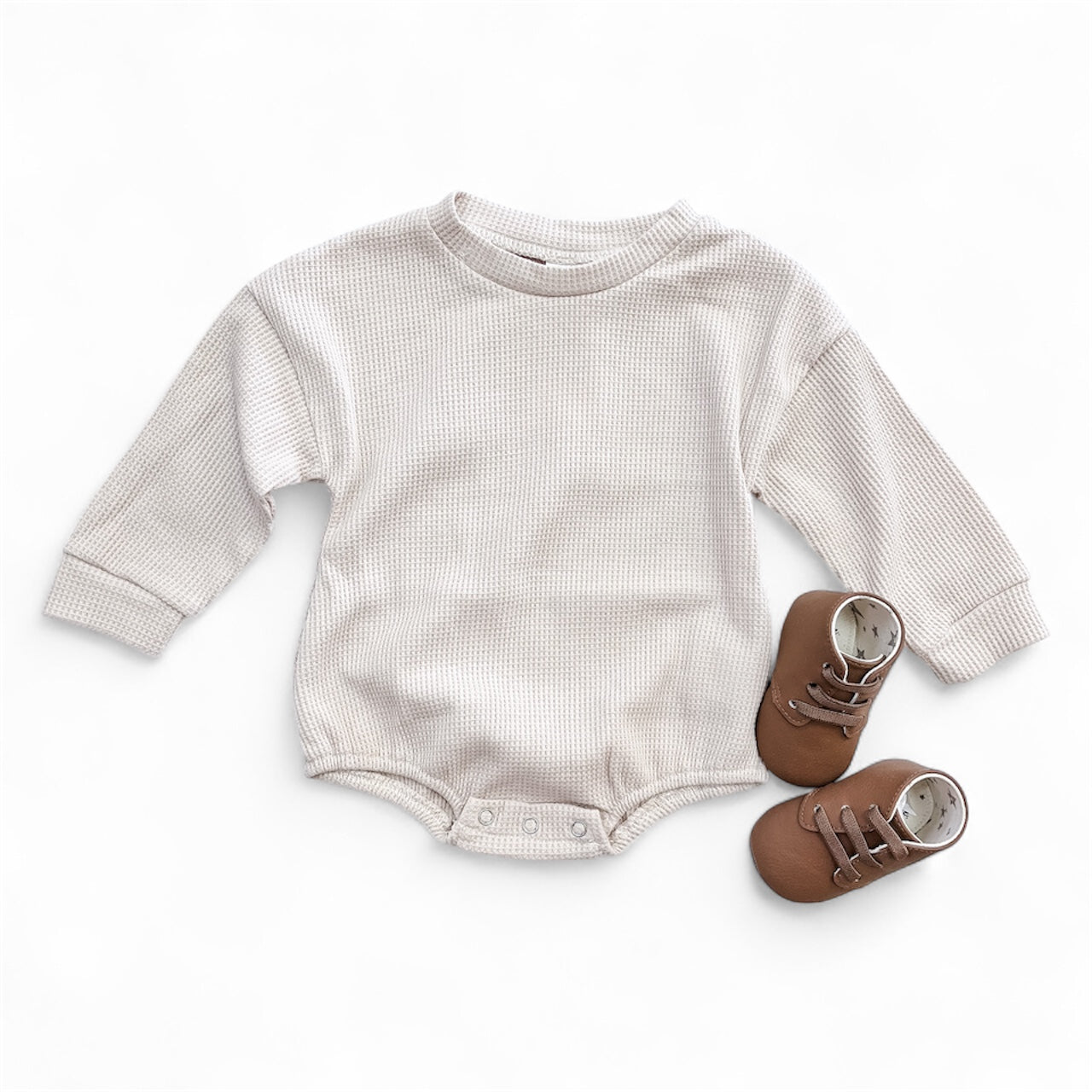 The Tiny Tot Bundles Neutral Nest Bubble, a beige long-sleeve baby onesie with snap buttons at the bottom, is laid flat beside small brown baby shoes on a white background.