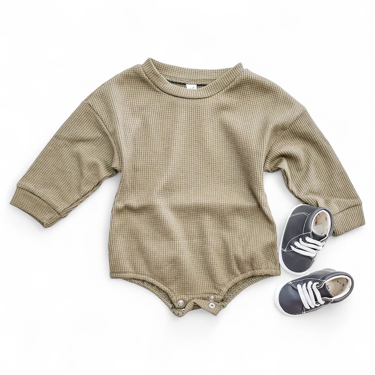 The Sage Wood Bubble by Tiny Tot Bundles, a beige long-sleeve baby romper with bottom snaps, is laid flat beside small gray baby sneakers with white laces on a white background.