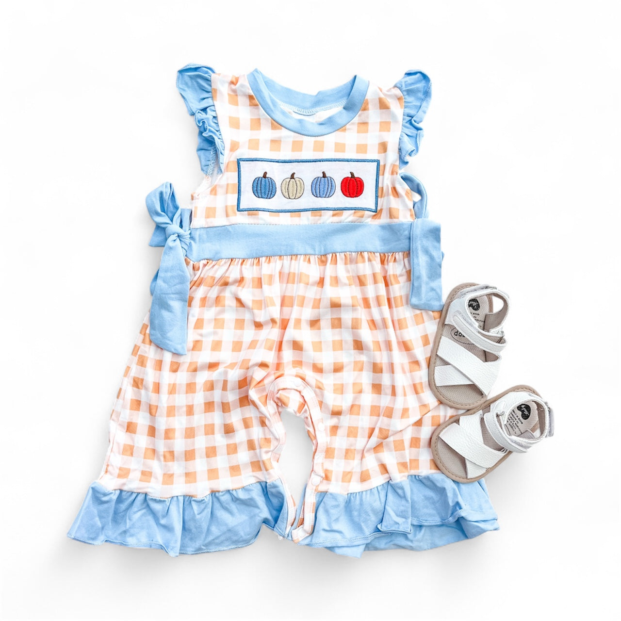 The Pumpkin Parade Romper by Tiny Tot Bundles features an orange and white gingham pattern, blue ruffles, and three pumpkin appliques, paired with white baby sandals on a white background.