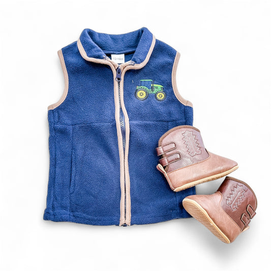 The Tiny Tot Bundles Little Tractor Vest, blue with light beige trim and a green tractor patch on the chest, is shown beside brown toddler cowboy boots featuring decorative stitching.