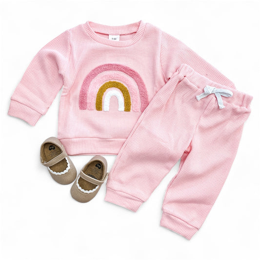The Chasing Rainbows Lounge Set by Tiny Tot Bundles includes a pink long-sleeve top with a rainbow design, matching pink pants with a white bow, and light brown baby shoes with white trim, all displayed on a white background.
