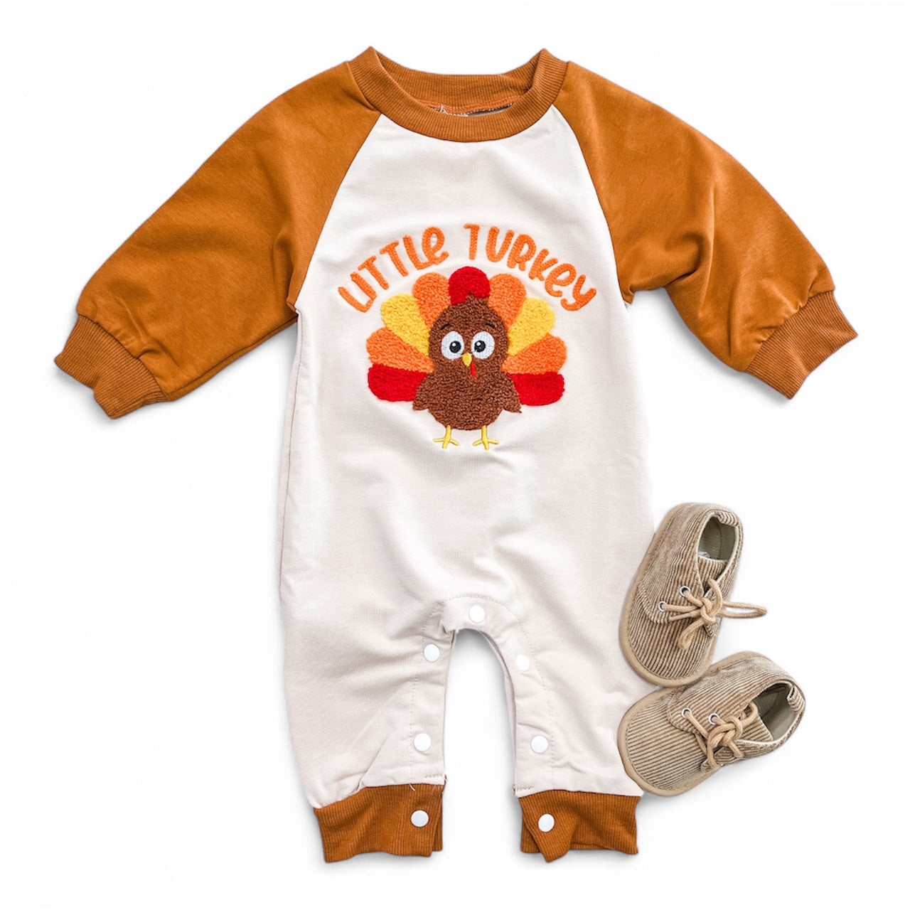 The Little Turkey Romper from Tiny Tot Bundles has brown sleeves and cuffs, a colorful turkey and “Little Turkey” on the front. Tan baby shoes are shown next to the romper on a white background.
