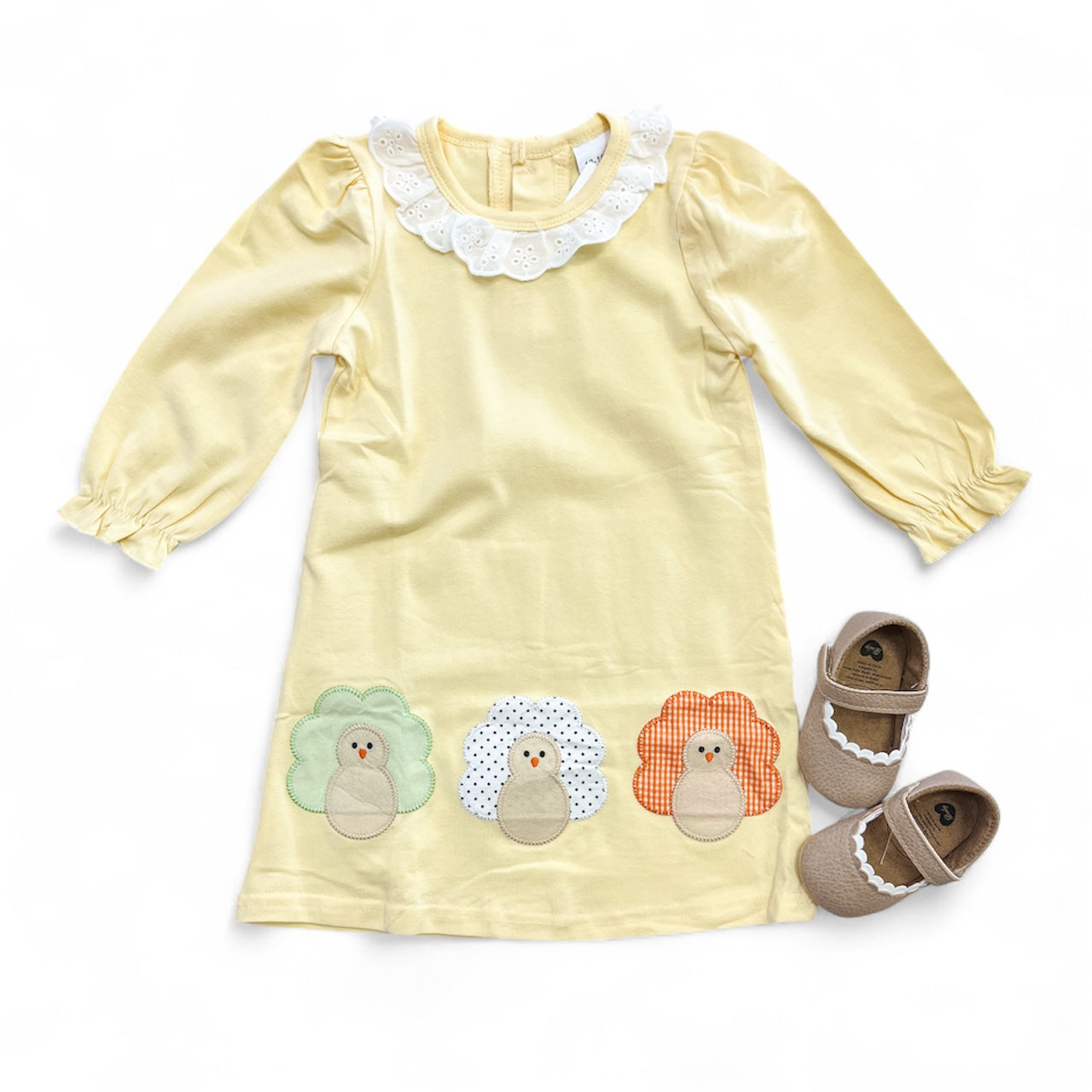 The Tiny Tot Bundles Turkey Day Parade Dress is a pale yellow long-sleeve dress with a white scalloped collar and three colorful turkey appliqués at the hem, shown with brown Mary Jane shoes on a white background.