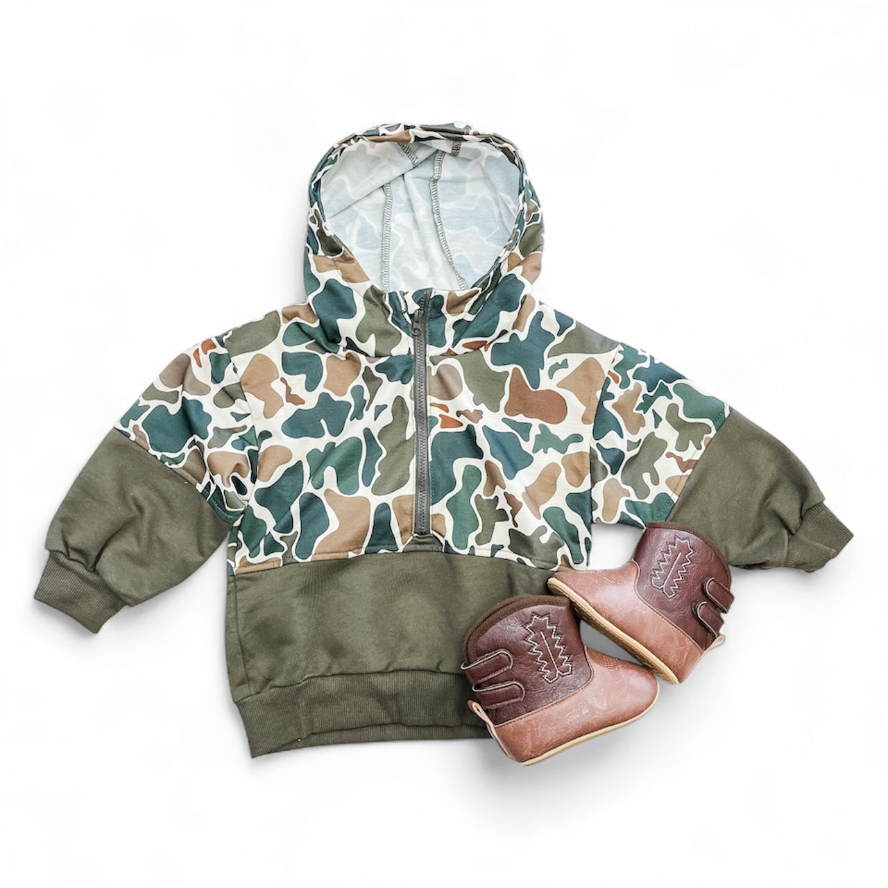 The Tiny Tot Bundles Mini Maverick Hoodie, featuring a green, brown, and beige camouflage pattern, is laid flat beside a pair of brown leather baby shoes with stitching details on a white background.