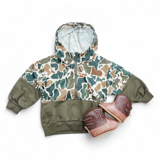 The Tiny Tot Bundles Mini Maverick Hoodie, featuring a green, brown, and beige camouflage pattern, is laid flat beside a pair of brown leather baby shoes with stitching details on a white background.