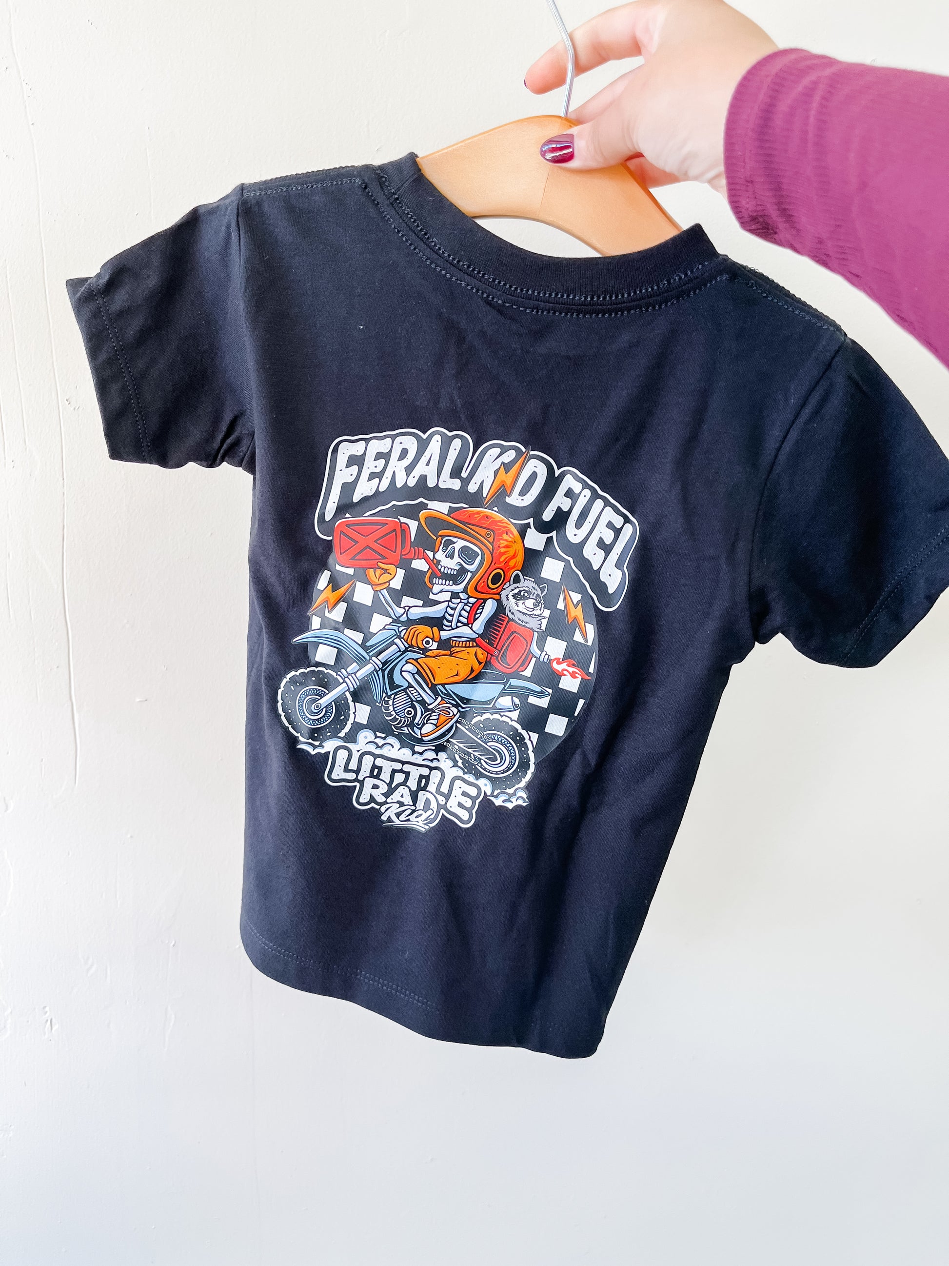 A hand holds the Little Rad Kid Feral Kid T-Shirt on a hanger. This small black tee features a colorful motocross rider graphic, "Feral Kid Fuel" and "Little Rebel" text, and a checkered flag pattern on the back.