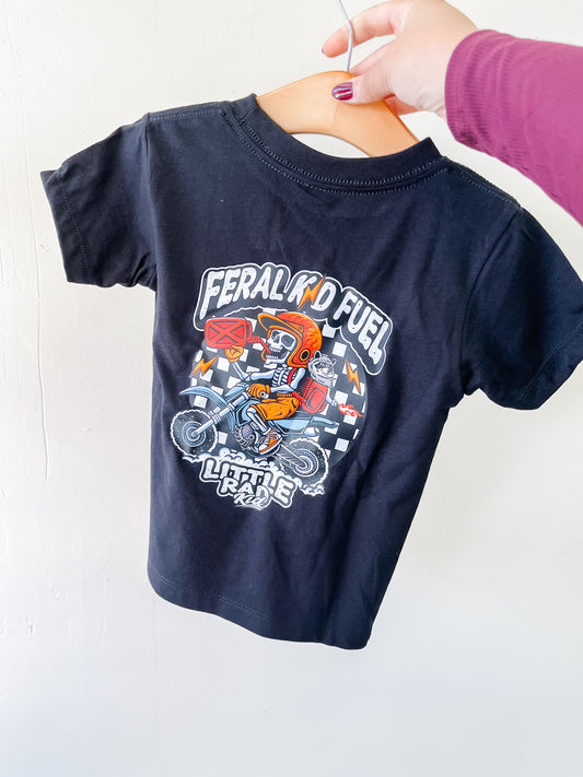 A hand holds the Little Rad Kid Feral Kid T-Shirt on a hanger. This small black tee features a colorful motocross rider graphic, "Feral Kid Fuel" and "Little Rebel" text, and a checkered flag pattern on the back.