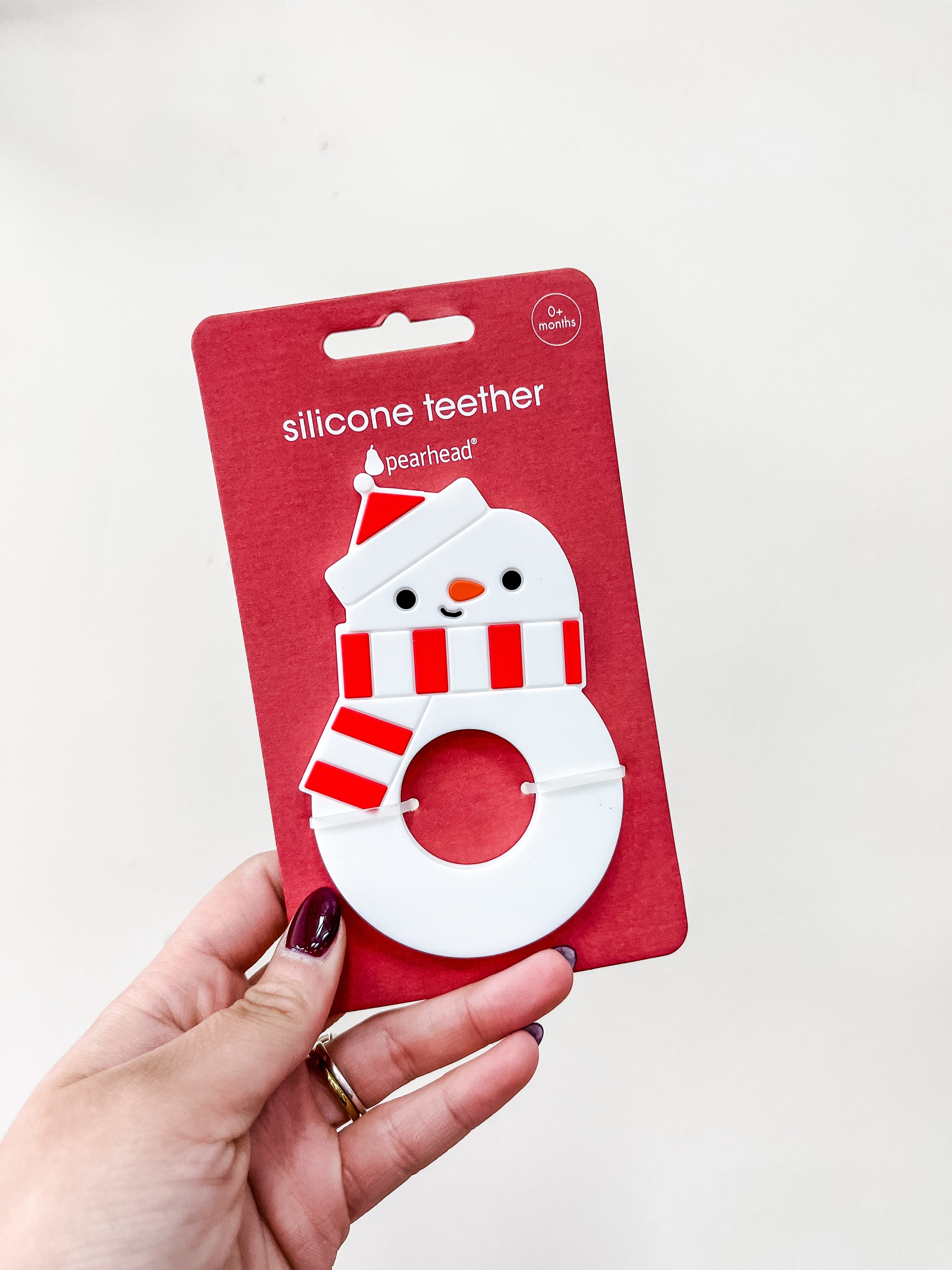 A hand holds a PearHead Snowman Silicone Christmas Baby Teether, shaped like a snowman with a red and white hat and scarf, packaged on a red card labeled “silicone teether” and “0+ months.”.