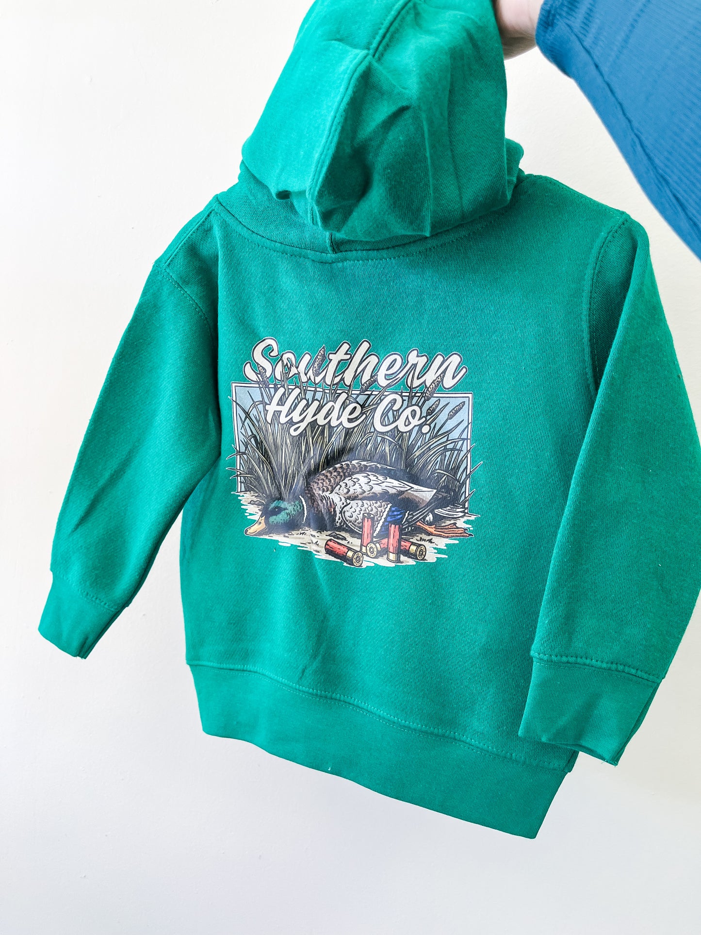 The Shell Shock Hoodie by Southern Hyde Co features a green design with a duck, grass, shotgun shells, and "Southern Hyde Co." printed on the back. Shown held up by the hood.