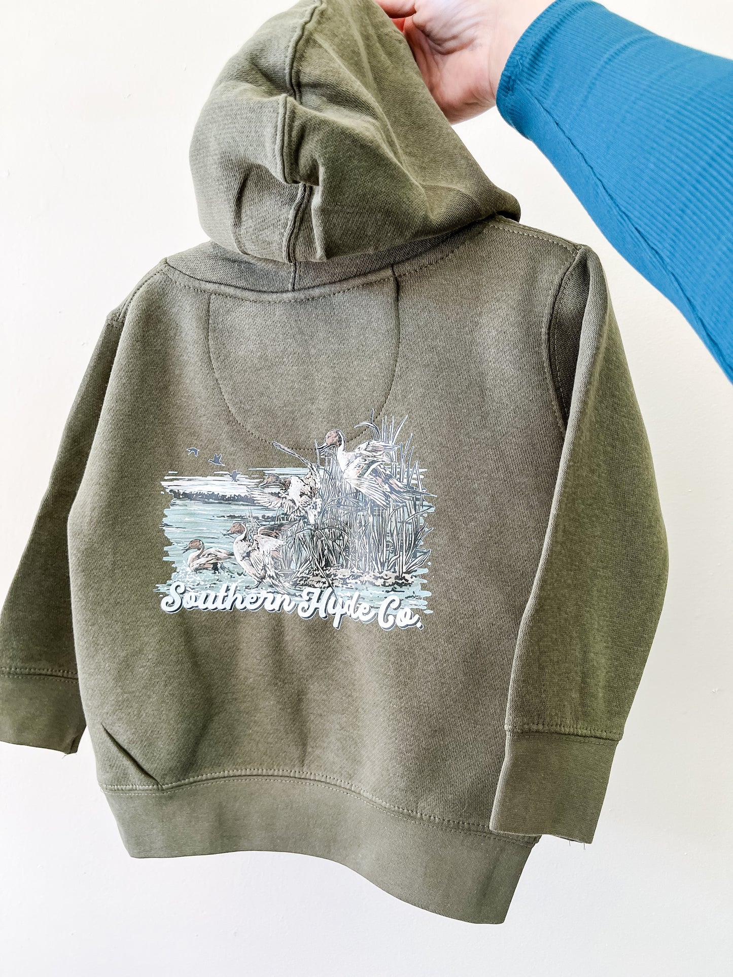 A hand holds up an olive green Pintails Skies Hoodie by Southern Hyde Co, featuring an outdoor scene with ducks and reeds on the back.